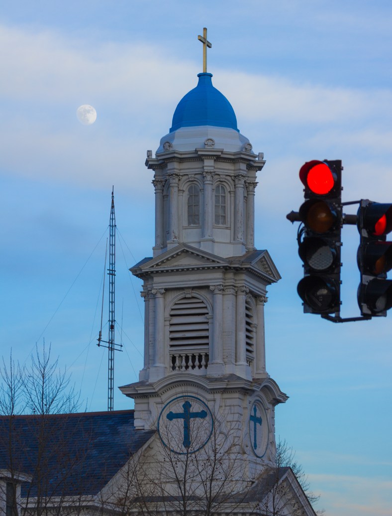 Behind two red lights at the intersection of Jason Street and Massachusetts Avenue, the Highrock Chruch, and behind that, the moon. January 13, 2014.